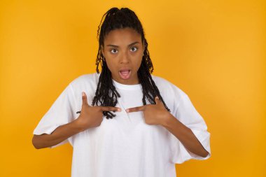Afro-Amerikalı kadın, şaşkın bir ifadeyle sarı arka planda kendine işaret ediyor.