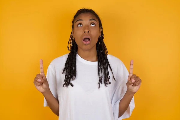 shocked african american woman against yellow background pointing fingers up, looking up with opened mouth