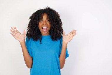 İyimser, kıvırcık saçlı, günlük mavi tişörtlü, beyaz arka planda duran Afrikalı genç bir kadın sevinçten avuçlarını kaldırıyor, birinden müthiş bir hediye almaktan mutlu, yüksek sesle bağırıyor, heyecanlı beyaz bir erkek çığlık atıyor..