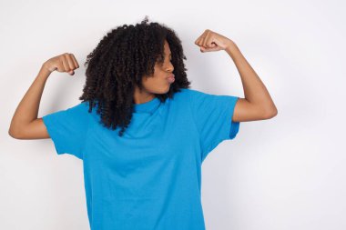 Kıvırcık saçlı, sıradan mavi gömlekli, beyaz arka planda duran genç Afrikalı kadın, gururla gülümseyen kol kaslarını gösteriyor. Fitness konsepti.