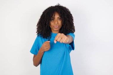 Kıvırcık saçlı, günlük mavi tişörtlü, beyaz arka planda duran, yumruklarını yumruklayan, saldırgan ve öfkeli bir kadın. Tehdit ve şiddet.