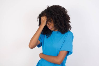 Kıvırcık saçlı, mavi gömlekli, beyaz arka planda duran üzgün ve yalnız bir Afrikalı kadın. Ağlıyor, 