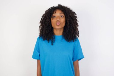 Hoş görünümlü, kıvırcık saçlı, sıradan mavi gömlekli, beyaz arka planda duran, dudaklarını büzen, kameraya yeşil gözlü, kız arkadaşıyla eğlenen, insan yüz ifadeleri takınan bir Afrikalı kadın.