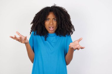 Hüsrana uğramış kıvırcık saçlı, sıradan mavi tişörtlü, beyaz arka planda duran genç Afrikalı kadın şaşkın ve tereddütlü hissediyor, omuz silkiyor, ağzı açık, ne yapacağını bilmiyor..