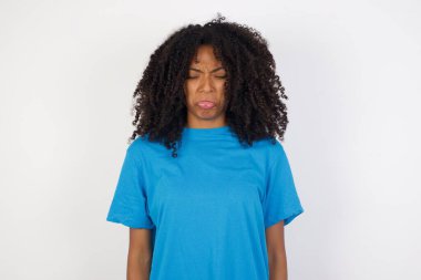 Kasvetli, reddedilmiş, kıvırcık saçlı, beyaz arka planda duran günlük mavi gömlekli genç Afrikalı kadının sorunları ve zorlukları var, alt dudak kıvrımları var ve umutsuzluk içinde gözlerini kapatıyor, depresyonda. 