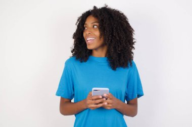 Çekici, kıvırcık saçlı, mavi tişörtlü, beyaz arka planda duran çekici, tatlı, güzel kız arkadaş kendinden geçmiş, sosyal medyada bir şeyler gören Afrikalı genç bir kadının fotosu. 