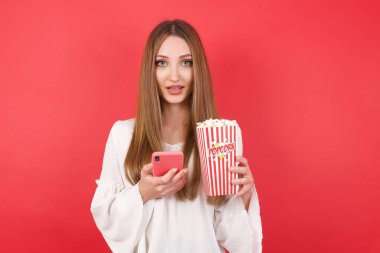 Kırmızı stüdyo arka planında poz veren patlamış mısır ve akıllı telefonlu güzel bir kadın.