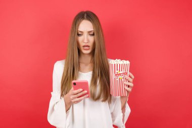 Kırmızı stüdyo arka planında poz veren patlamış mısır ve akıllı telefonlu güzel bir kadın.