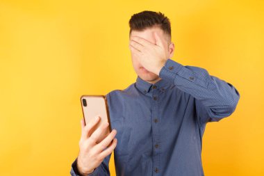 Stüdyoda ağlayan adam sarı arka planda, yüzünü eliyle kapatıyor ve cep telefonunu tutuyor. 