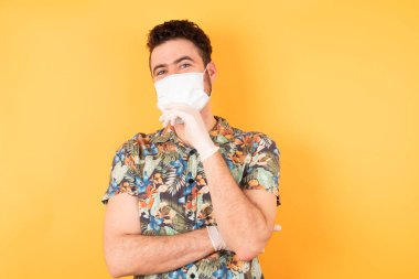 Koruyucu maskeli yakışıklı genç adam ellerini kısmen çapraz yapıyor ve elleri çenenin altında, zevkle kameraya bakıyor. 