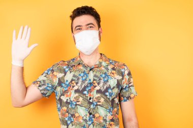 Koruyucu maskeli yakışıklı genç adam el sallıyor. Mutlu, dostane hoş geldin jesti..