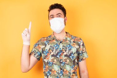 Koruyucu maskeli genç adam parmağını kaldırıyor. Fikri var ve sarı arka planda poz veriyor..