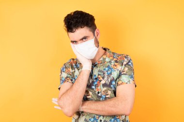 Koruyucu maskeli genç adam el ele tutuşurken diğer eli çapraz tutarak, yorgun ve hasta görünüyor. 