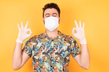 Koruyucu maskeli, gözleri kapalı genç ve yakışıklı bir adam parmaklarıyla meditasyon hareketi yapıyor. Yoga konsepti.