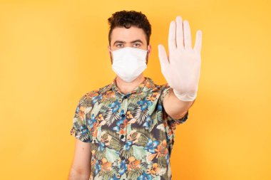 Koruyucu maskeli genç yakışıklı, avuç içiyle hareket etmeyi kesiyor. Sarı arkaplan üzerinde izole edilmiş yüz üzerinde negatif ve ciddi jest içeren uyarı ifadesi.