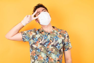 Koruyucu maskeli yakışıklı genç adam barış sembolü yapıyor. Yüzünde parmaklar, gülümseyerek zaferi gösteriyor.