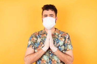 Sarı stüdyoda tıbbi maskeli beyaz adam, üzgün bir yüzle yalvarıyor ve dua ediyor.