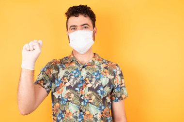 Sarı stüdyoda tıbbi maskeli beyaz bir adam. Eldivenli yumruğunu gösteriyor. 