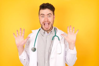 İyimser doktor sevinçten avuçlarını kaldırır, harika haberler aldığına sevinir, yüksek sesle bağırır, tıbbi üniforma giyer, sarı arka planda izole olur. Heyecanlı beyaz erkek çığlıkları.