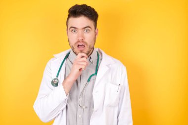 Gergin, kafası karışık bir doktorun portresi, şaşkınlıktan ağzı açılır, ani haberlere tepki verir, sarı arka planda izole bir şekilde tıbbi üniforma giyer. İnsanlar ve yüz ifadeleri.