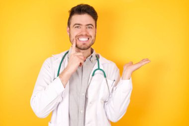 Yakışıklı, komik bir adamın elinde yeni bir ürünün fotoğrafı. Harika bir öneri. Sarı arka planda tek başına duran tıbbi üniforma giyiyor..