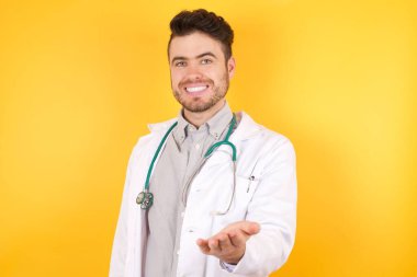 Sarı arka planda gülümseyen doktor mutlu bir şekilde avuç içi yardım ve kabullenme sunuyor..