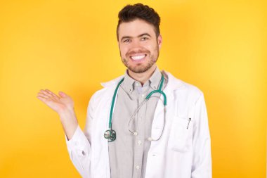 Genç beyaz doktor, izole edilmiş sarı arka planda tıbbi üniforma giyiyor, gülümsüyor ve avucunun içi kameraya bakıyor..