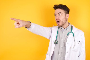 Sarı arka planda tıp üniforması giyen yakışıklı beyaz bir doktor. Parmakla işaret ederek şaşırdı, ağzını açtı. Şaşırdı, ön tarafta bir şey vardı..