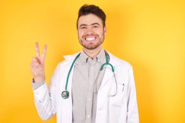 Sağlık üniforması giyen, sarı arka planda duran yakışıklı, beyaz bir doktor. Gülümseyerek kameraya göz kırparak zafer işareti yapıyor. İki numara..