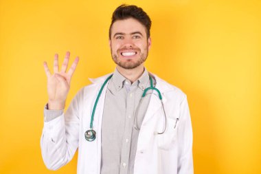Avrupalı yakışıklı doktor üniformasını giymiş sarı arka planda duruyor, dört numaralı parmaklarıyla gösteriyor ve işaret ediyor kendinden emin ve mutlu gülümsüyor..