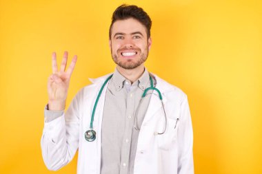 Genç Avrupalı yakışıklı doktor üniforması giyiyor. Sarı arka planda duruyor. Üç numaralı parmaklarını gösterip işaret ederken mutlu ve kendinden emin gülümsüyor..