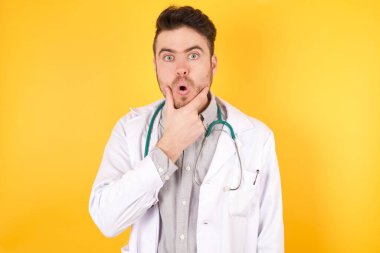 Sarı arka planda duran doktor üniforması giyen yakışıklı beyaz bir doktor. İnançsızlıktan, şaşkınlıktan ve elleri çenesinde şaşkınlıktan büyülenmiş bir ifade.