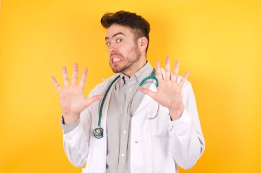 Sağlık üniforması giyen yakışıklı beyaz bir doktor, izole edilmiş sarı bir geçmişi var. Korku ifadelerinden korkuyor ve dehşete düşüyor. El hareketlerini durdur, şok içinde bağır. Panik konsepti.