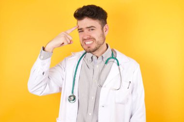 Tıbbi üniforma giyen yakışıklı beyaz bir doktor, izole edilmiş sarı arka planda duruyor, alnındaki sivilceden mutsuz olduğunu gösteriyor, siyah nokta enfeksiyonu. Cilt bakımı kavramı.