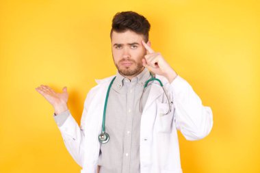 Tıbbi üniforma giyen yakışıklı beyaz bir doktor, izole edilmiş sarı arka planda duruyor. Avuç içinin açık olması, fotokopi alanını göstermesi ve alnını işaret etmesi onu rahatsız ediyor. Bunu bir düşün..