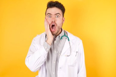 Şok olmuş beyaz doktor üniforması giyen yakışıklı adamın yatay portresi beklenmedik haberler karşısında şaşkınlığa uğramış, sarı arka plan karşısında modeller. Yüz ifadeleri kavramı.