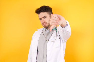 Sağlık üniforması giyen yakışıklı beyaz bir doktor kızgın, sinirli, hayal kırıklığına uğramış ya da canı sıkkın sarı duvara ciddi ciddi bakıyor.