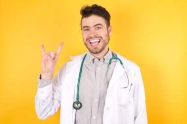 Sağlık üniforması giyen yakışıklı beyaz bir doktor kameraya gülümsüyor. Sarı duvara yaslanmış. En sevdiği konsere gitmeye hazır..