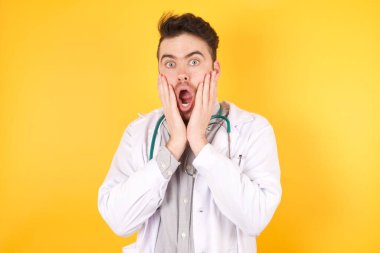 Ürkmüş beyaz doktor üniforması giyen bir stüdyo fotoğrafı dükkandaki fiyatlardan şok olmuş sarı arka planda izole edilmiş bir şey almak için para sıkıntısı çekiyor. İnsanlar ve insan duyguları kavramı