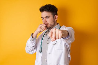 Güçlü ve kararlı doktor üniforması giymiş bir adamın portresi. Havayı yumrukluyor ve kameraya özgüvenle bakıyor. Kadın mücadelesi, savaşçı ruhu. Stüdyo çekimi sarı arkaplanda izole edildi