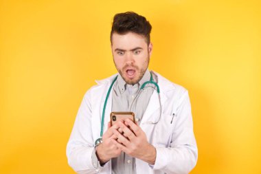 Tıbbi üniforma giyen beyaz doktor portresi, sosyal medya haberlerini ya da önemli e-postaları okuyarak sarı arka planda duran akıllı telefon kullanın..