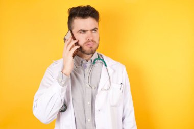 Üzgün, koyu saçlı, beyaz, doktor üniformalı adam sarı arka planda akıllı telefondan konuşuyor. İletişim konsepti.