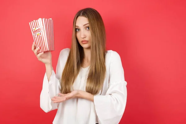 Young beautiful woman with popcorn pointing aside with hand showing something strange and saying: I don\'t know what is this. Advertisement concept.