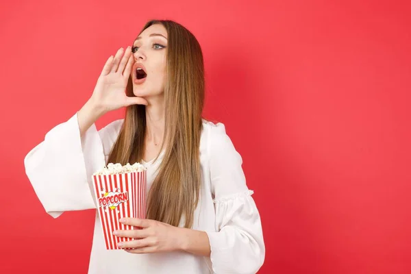 Young beautiful woman with popcorn shouting and screaming loud to side with hand on mouth. Communication concept.