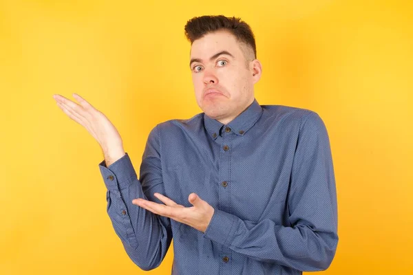 handsome young businessman pointing aside with both hands showing something strange and saying: I don\'t know what is this. Standing against gray background. Advertisement concept.
