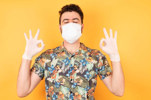Koruyucu maskeli, gözleri kapalı genç ve yakışıklı bir adam parmaklarıyla meditasyon hareketi yapıyor. Yoga konsepti.
