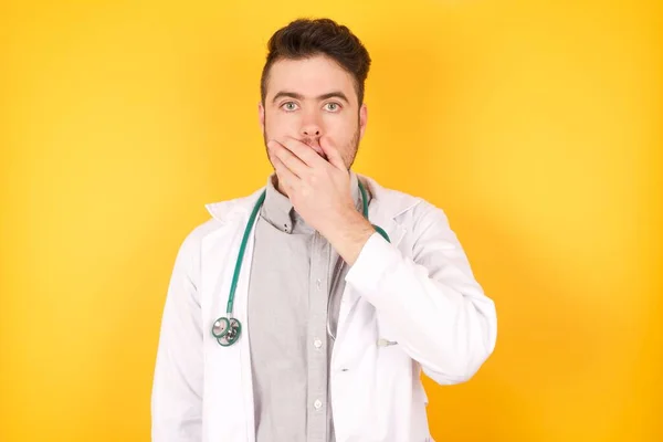 Emotional Caucasian doctor man gasps from astonishment, covers opened mouth with palm, looks shocked at camera, wears medical uniform standing over yellow background. Reaction concept.