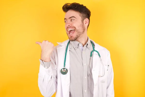 若いですハンサムな白人医師男身に着けています医療制服上の黄色隔離された背景 笑顔で幸せな顔見ますと指で側面を指していますアップ — ストック写真