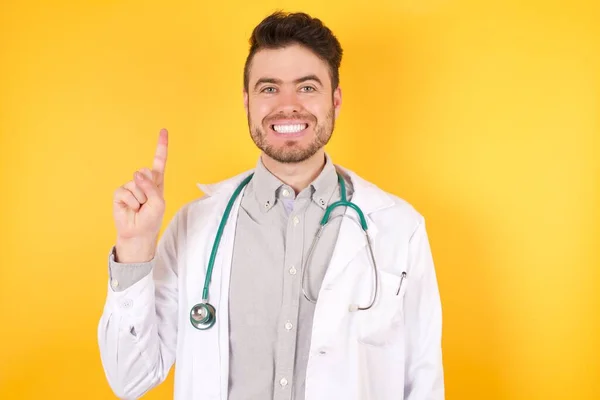 身穿医疗制服的年轻的欧洲英俊医生站在孤立的黄色背景之上 带着第一指尖出现 面带微笑 自信而快乐 — 图库照片