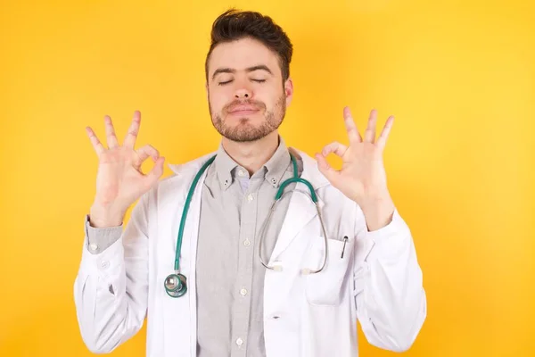 Beyaz arka planda tıbbi üniforma giyen yakışıklı beyaz doktor rahatla ve parmaklarıyla meditasyon hareketi yaparken gözleri kapalı gülümse. Yoga konsepti.